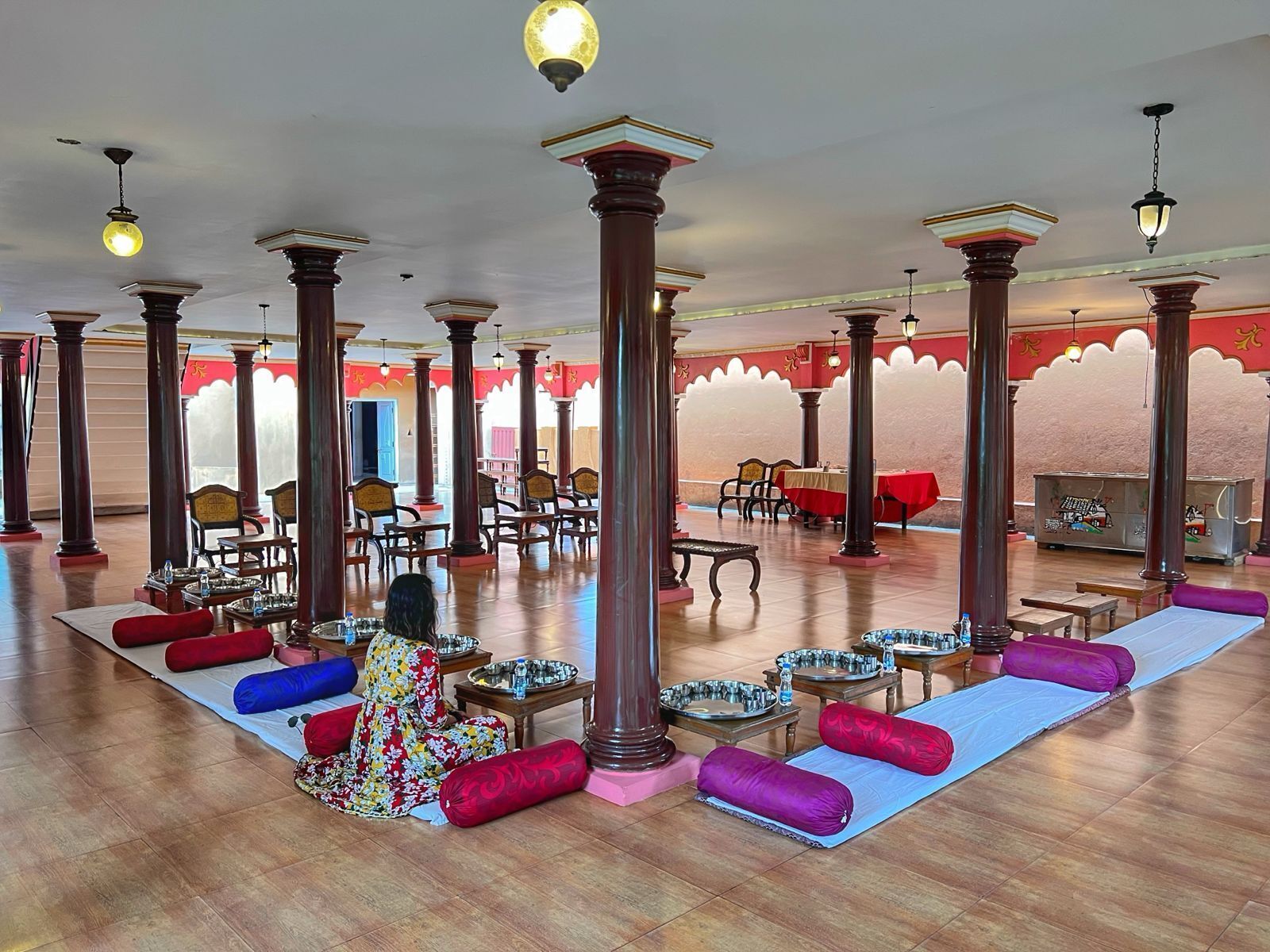 A spacious hall with high wooden pillars, multiple tables with chairs, and a central area with a mat and cushions.