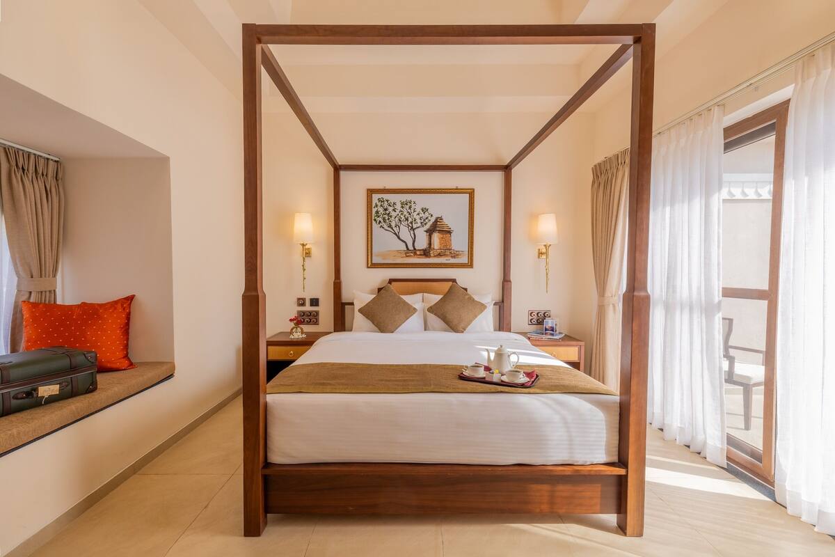 A four-poster bed with a white bedspread, a brown throw blanket, and a tray with a teapot and cups. There is a painting on the wall above the bed, and a window with curtains on the right side.
