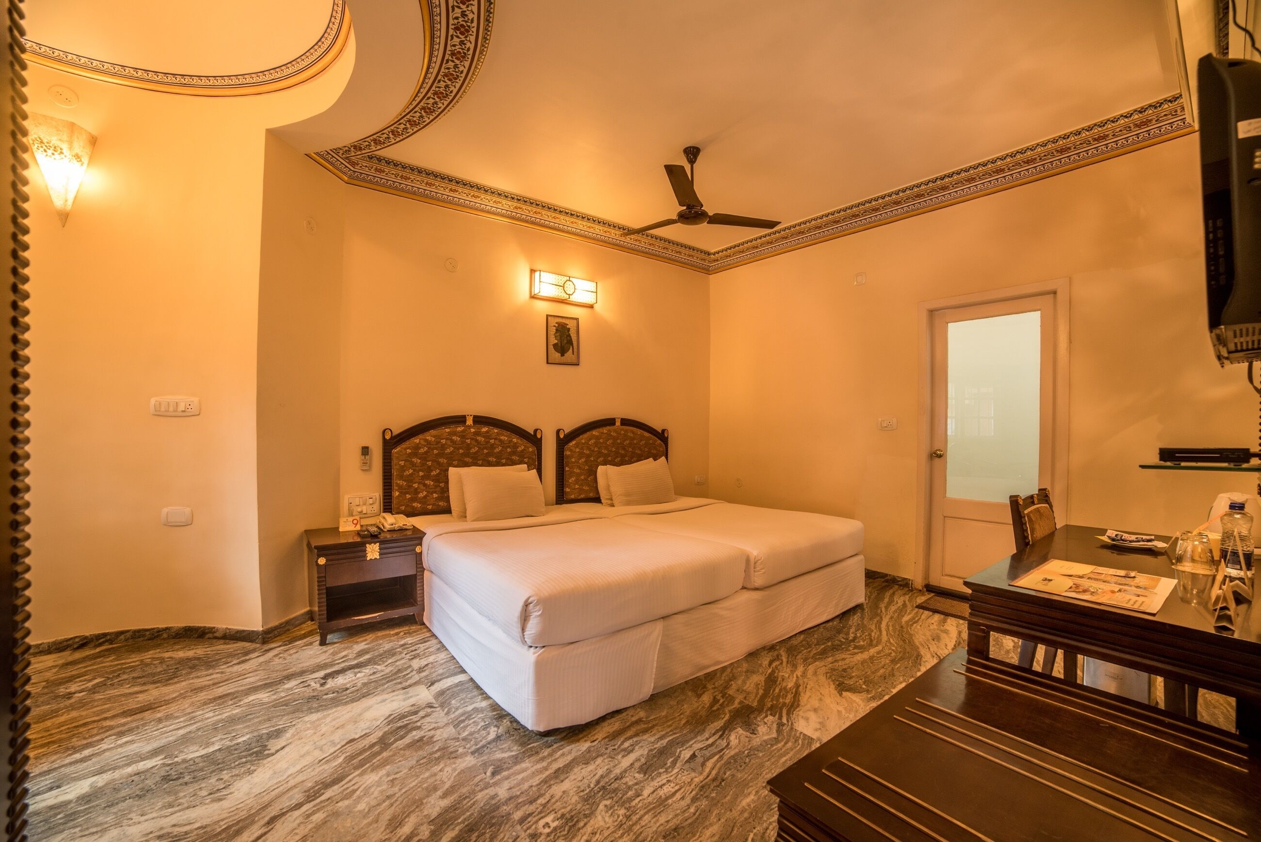 A hotel room with a bed, a desk, a chair, a nightstand, a ceiling fan, a wall-mounted light, and a door.