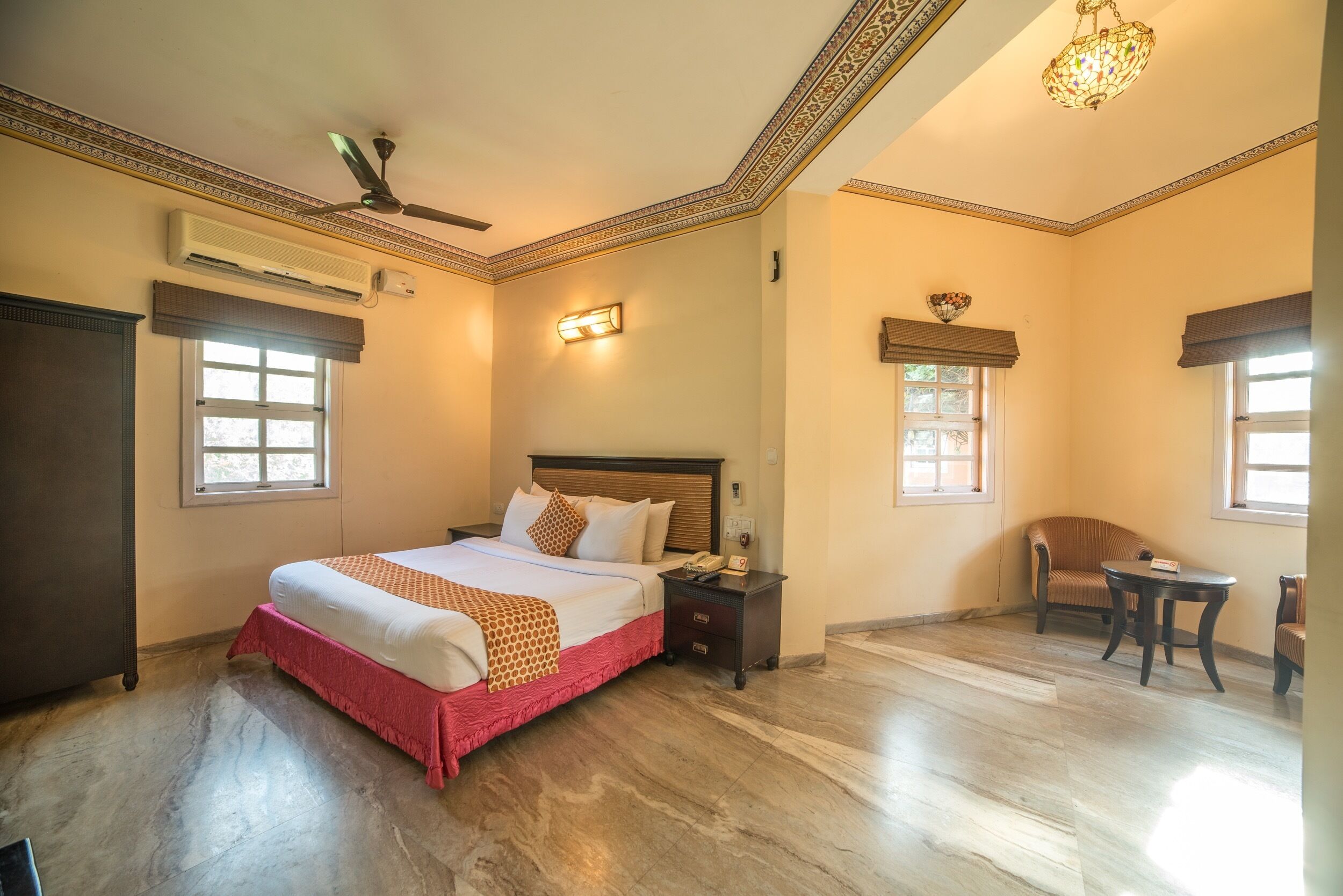 A hotel room with a large bed, a nightstand, a chair, and a small table. There is a ceiling fan, a chandelier, and a window with a view.
