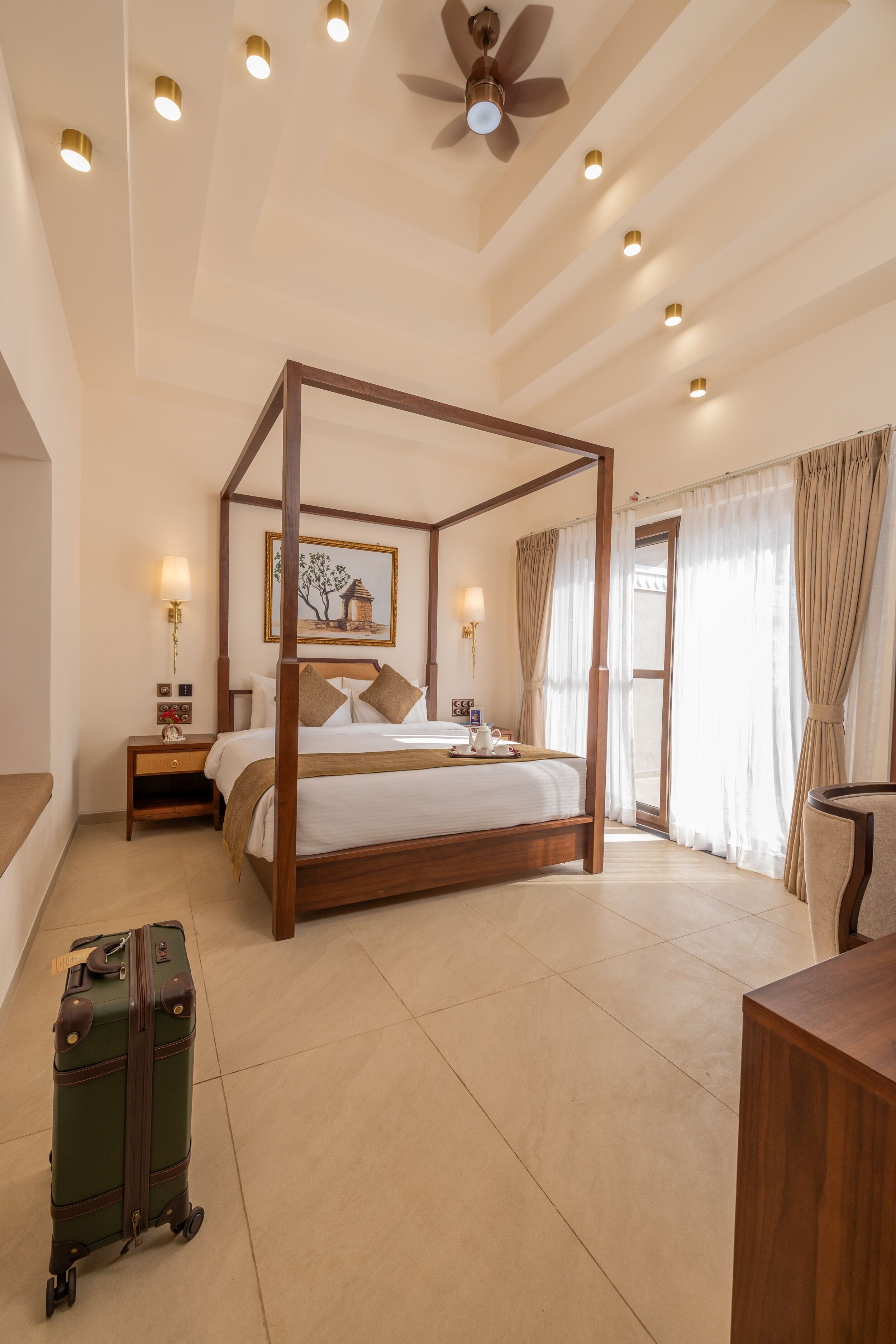 A four-poster bed with a wooden frame, a nightstand with a lamp, a suitcase, and a wooden desk.