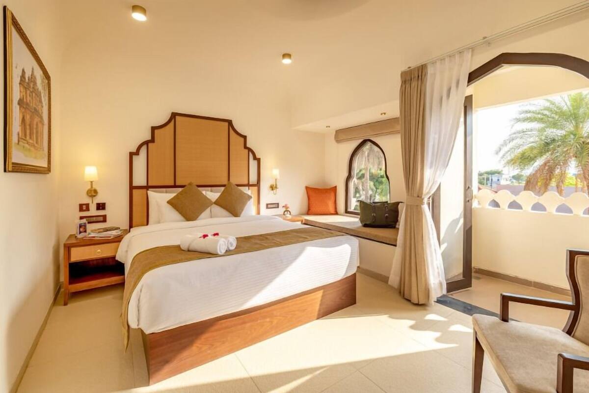 A hotel room with a large bed, a nightstand, a window with curtains, and a view of palm trees.