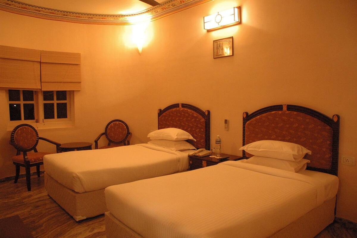 A hotel room with two single beds, a small table, two chairs, a window with blinds, and a wall-mounted light fixture.