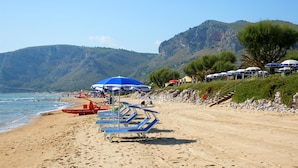 A beach with rows of blue lounge chairs and umbrellas, a lifeguard tower, and a mountainous backdrop. - Hotel Villaggio Torre San Vito (Gaeta)