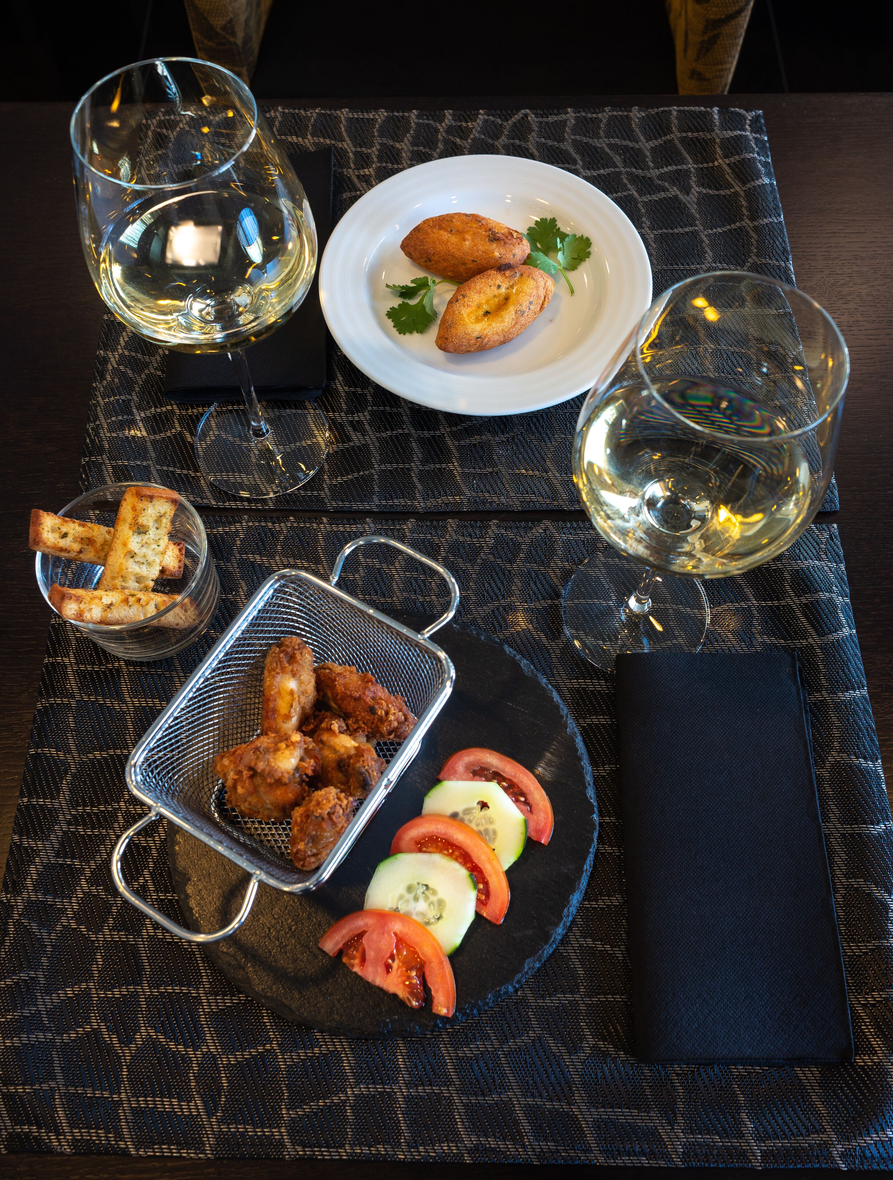 A dining table with a plate of food, a basket of fried chicken, a glass of white wine, and a glass of water.