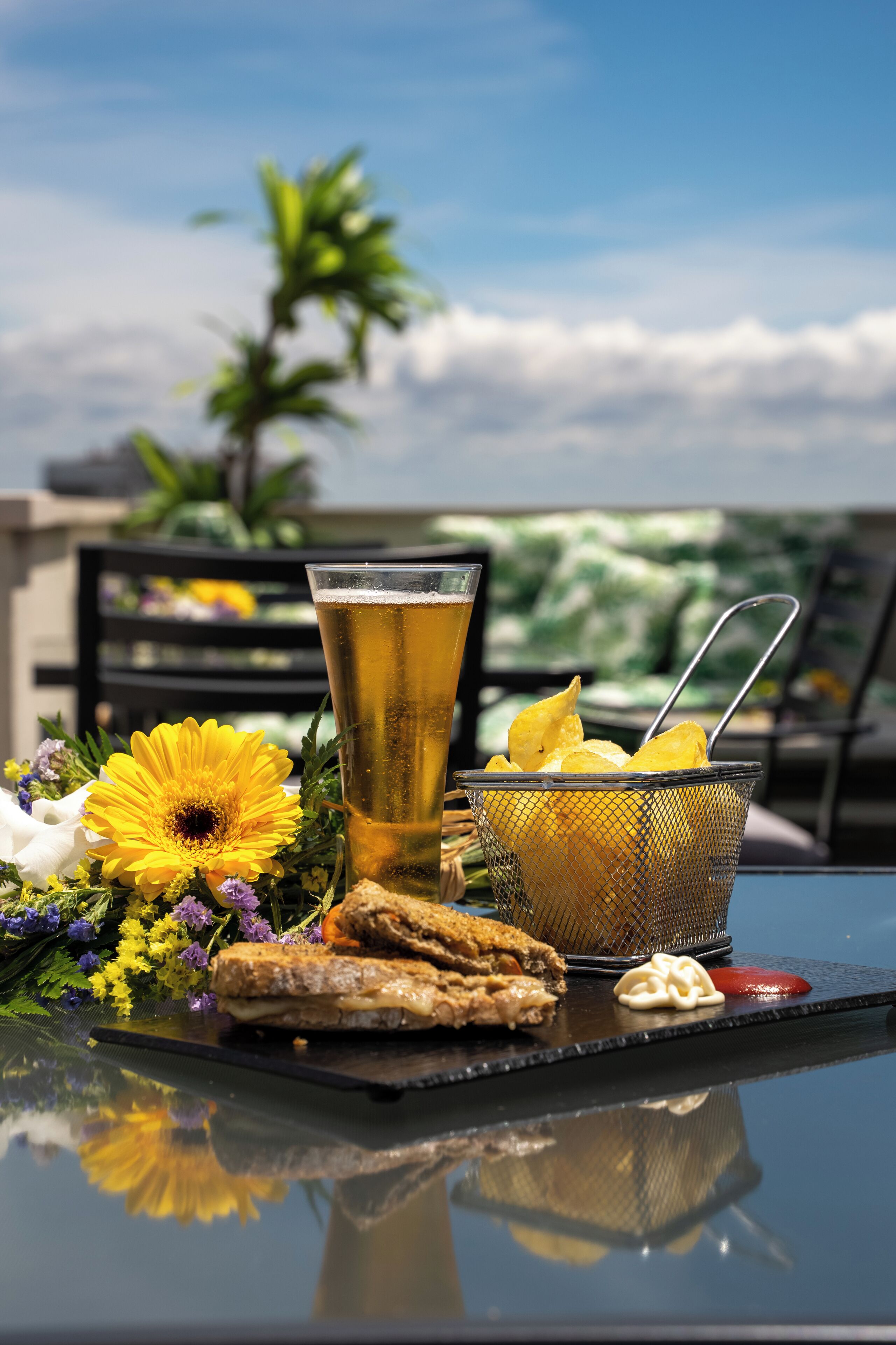 Ett bord med ett glas öl, en korg med chips, en smörgås och en liten skål med sås, placerat framför en bakgrund av blommor och en klar himmel.