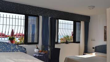 Habitación de hotel con dos camas, una mesa de comedor con frutero y vista al mar a través de ventanas grandes con cortinas.