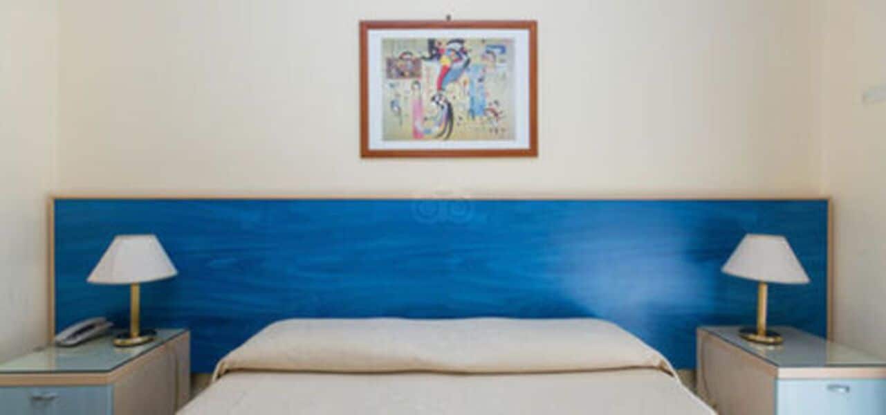 Une chambre à coucher comprenant un lit, deux tables de chevet, une commode, une lampe et une œuvre d’art encadrée accrochée au mur.