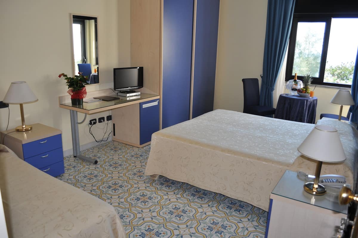 A hotel room with a bed, a desk with a computer, a chair, a small table with a fruit bowl, and a window with curtains.