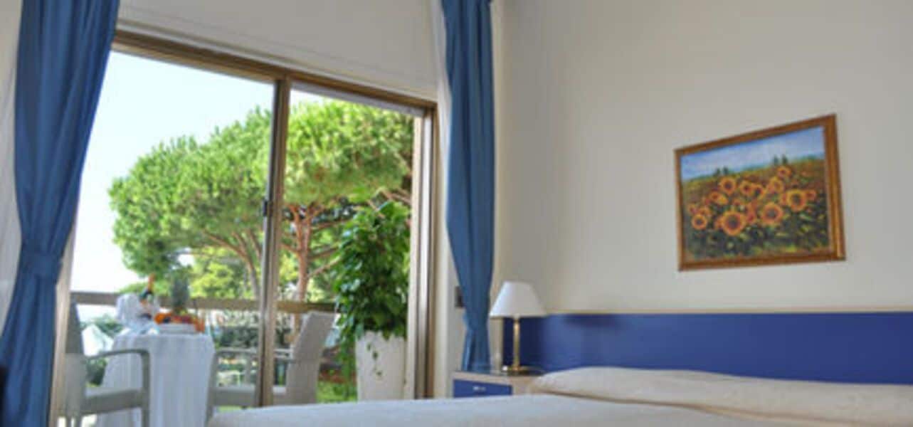 Quarto de hotel com cama, mesa e cadeira, janela com cortinas e um quadro na parede.