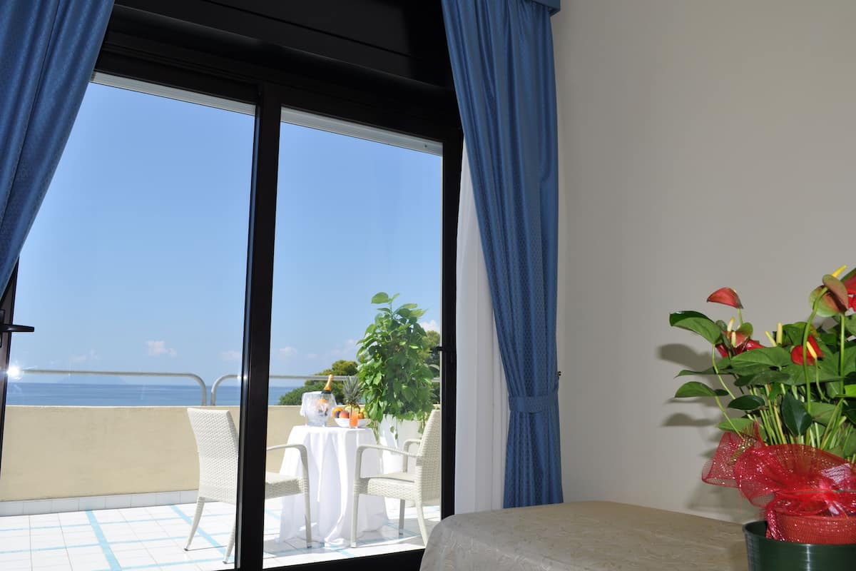 A room with a large window, a bed, a table with a plant, and a view of the sea.