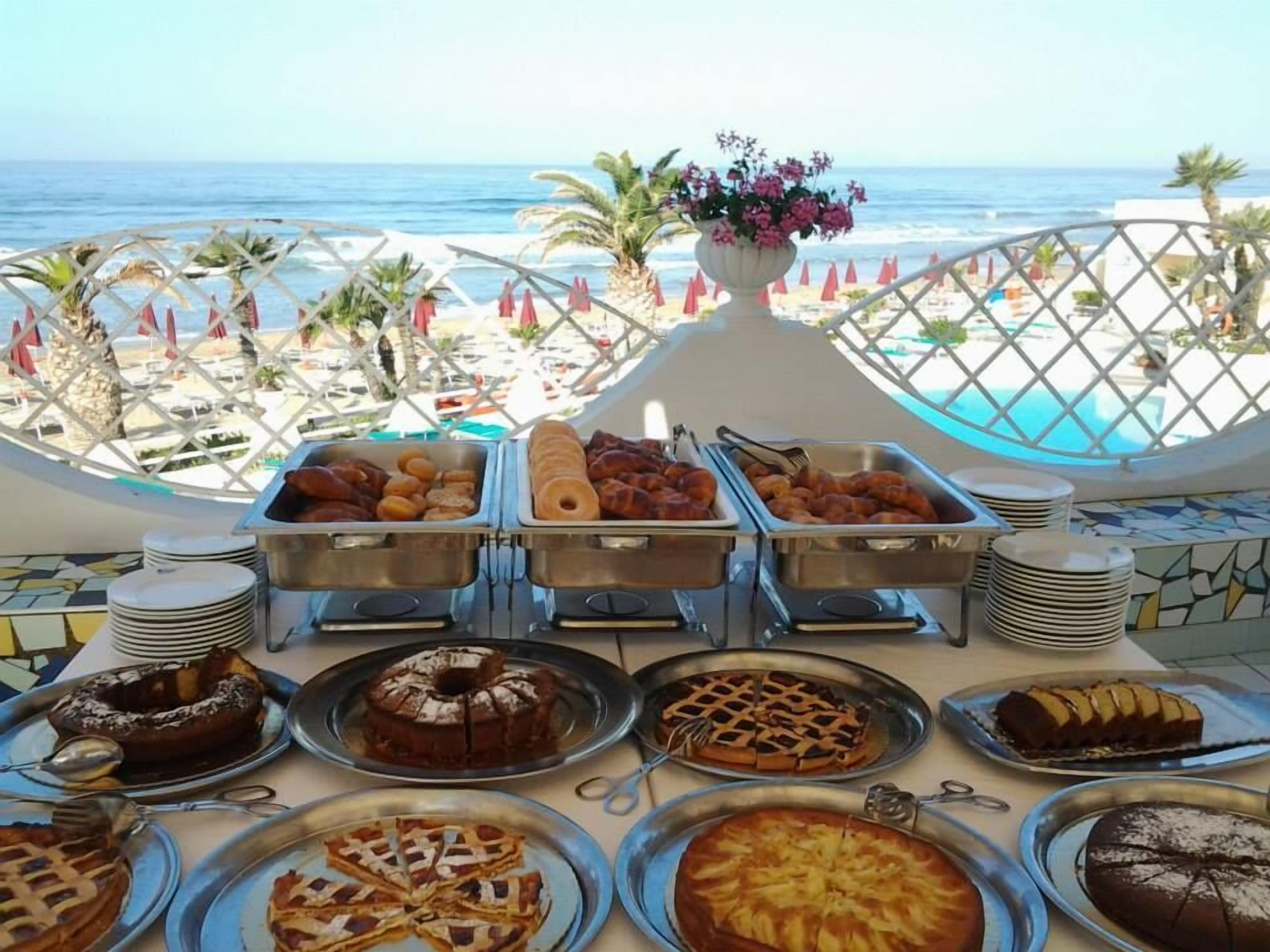 A buffet table with various desserts and pastries, overlooking a beach with umbrellas and palm trees.
