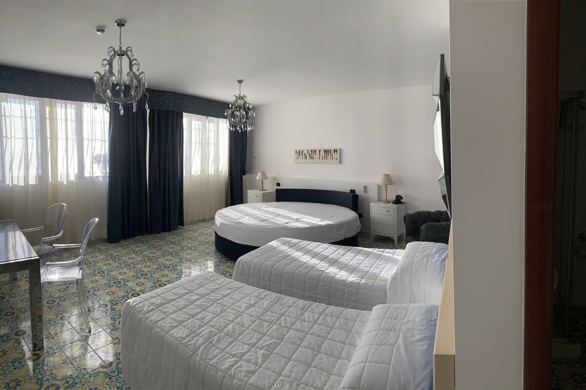 A hotel room with two beds, a chandelier, a desk with chairs, and a large window with curtains.