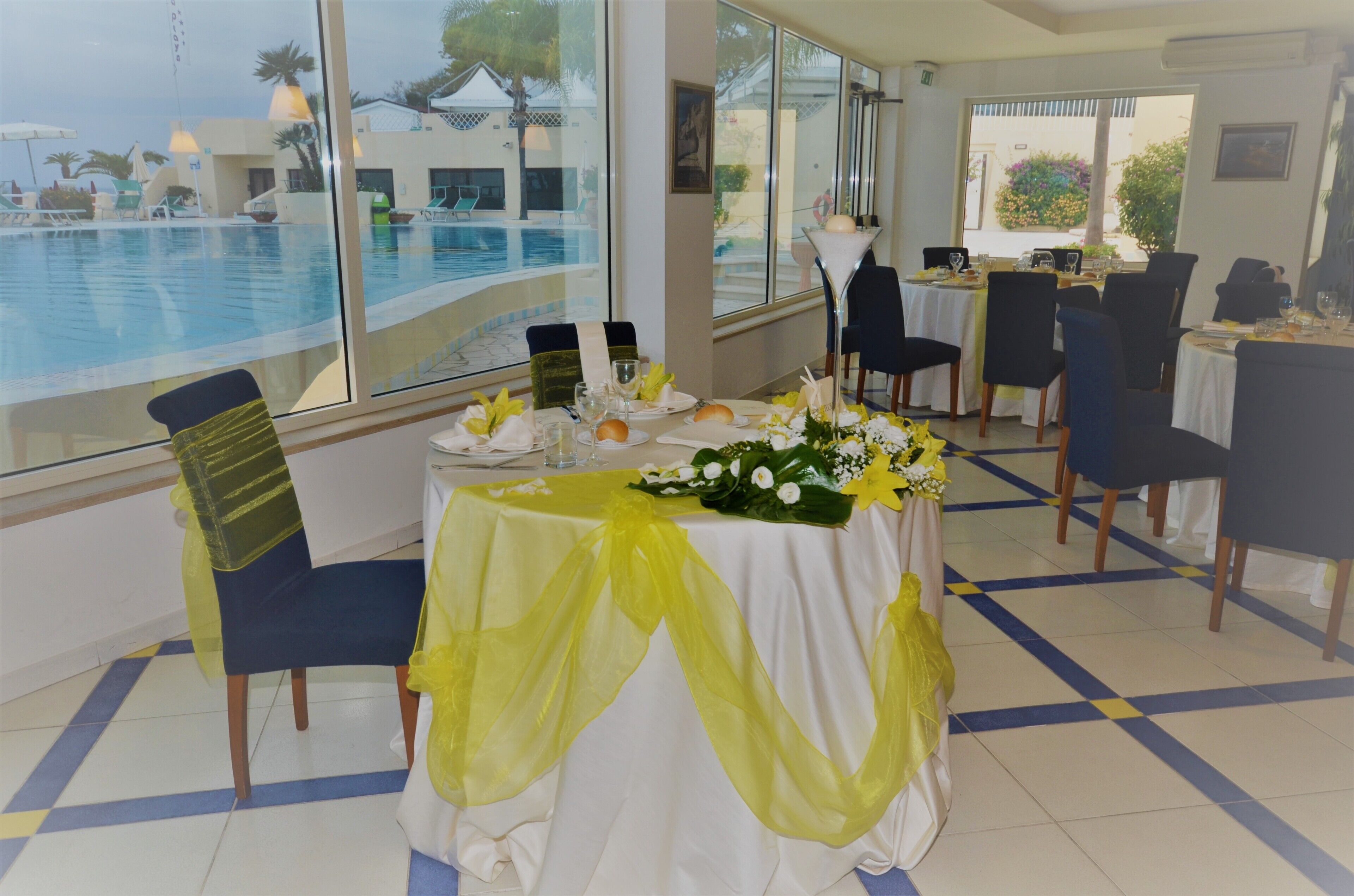 A dining area with a view of a swimming pool, tables set with white tablecloths and yellow accents, and chairs with green and blue upholstery.