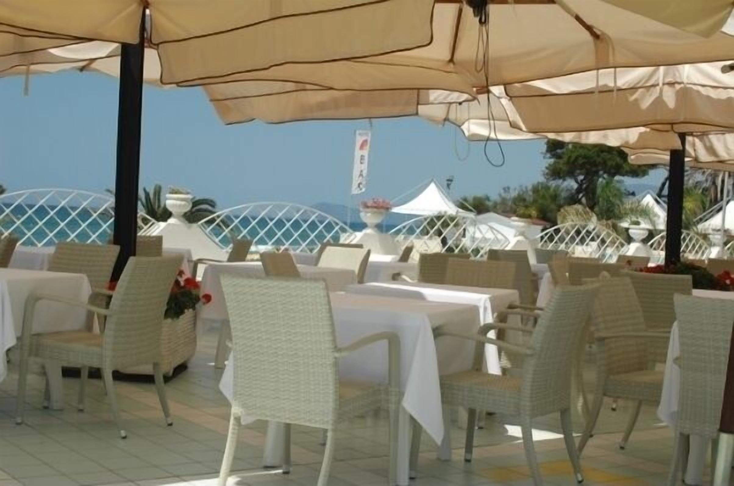 An outdoor dining area with white wicker chairs and tables, covered by beige umbrellas, overlooking a scenic view.