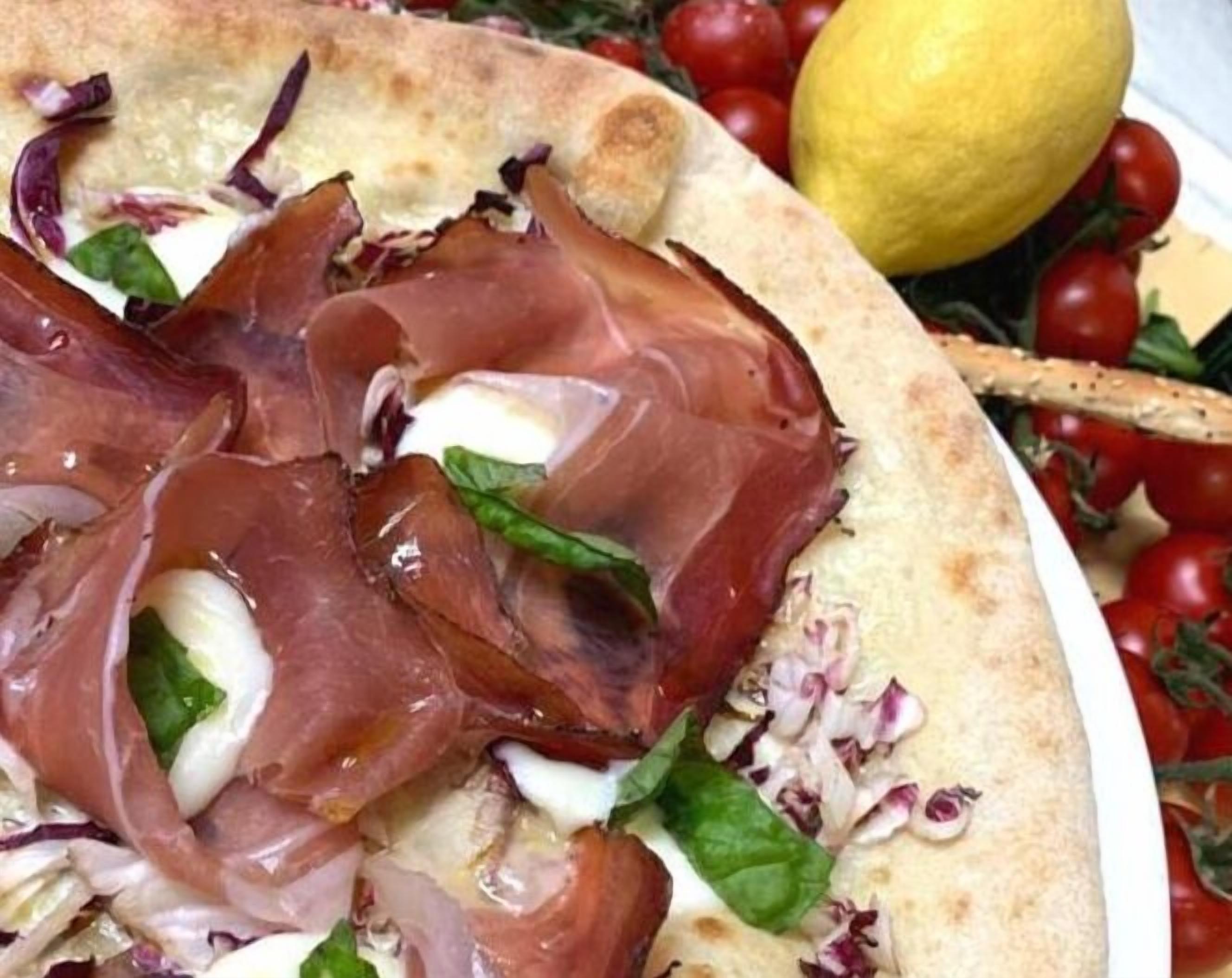 Pizza com presunto, mussarela e rúcula em um prato branco, acompanhada de tomates cereja e um limão.