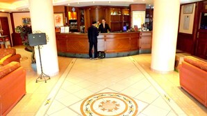 Un vestíbulo de hotel con una mesa de recepción, dos recepcionistas, un reloj y un diseño decorativo en el suelo.