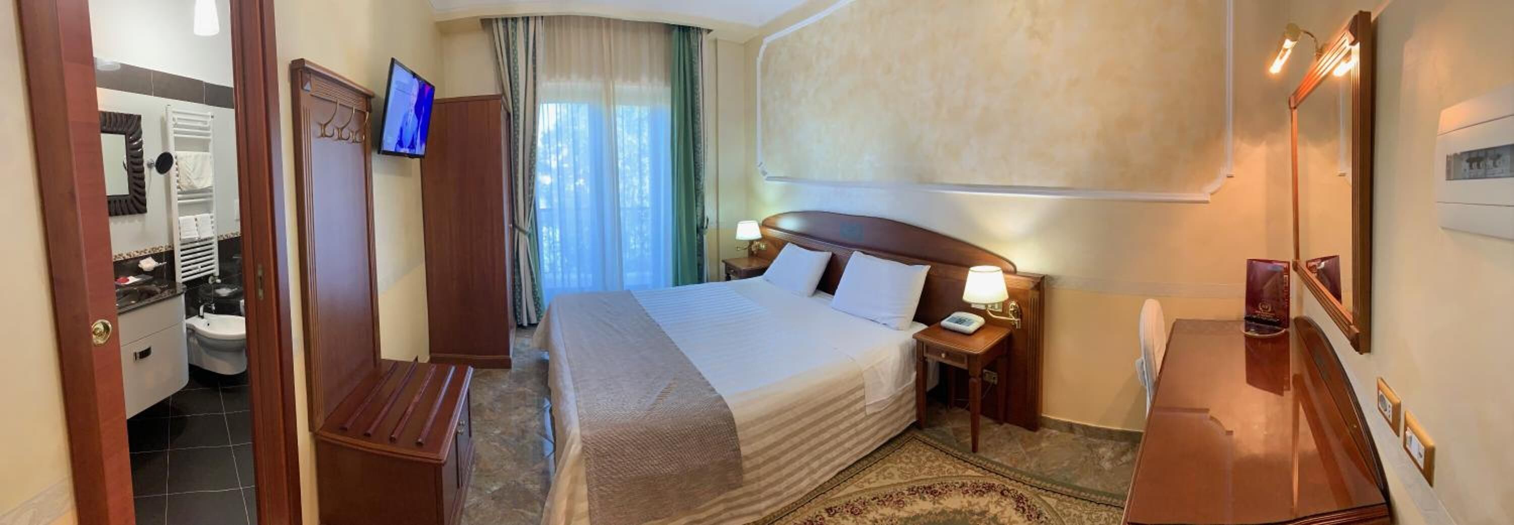 Una habitación de hotel con cama, mesitas de noche, televisión, espejo y baño con inodoro y lavamanos.