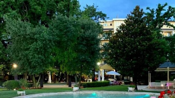 Un jardín ajardinado con una fuente, rodeado de árboles y un edificio al fondo.