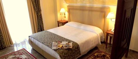 A hotel room with a bed, two bedside tables, a nightstand, a window with curtains, and a door.