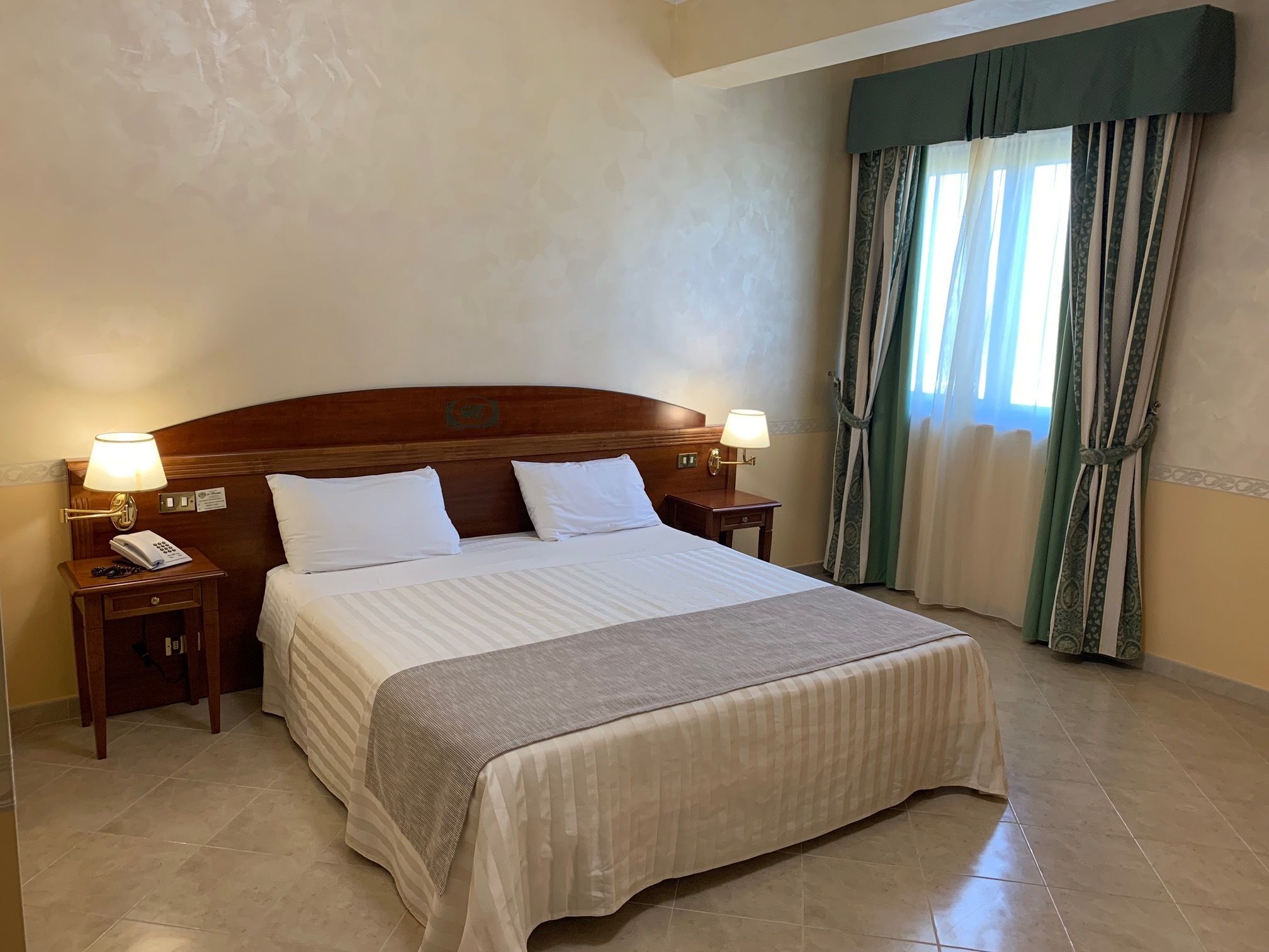 A hotel room with a large bed, two bedside tables with lamps, a telephone, and a window with curtains.