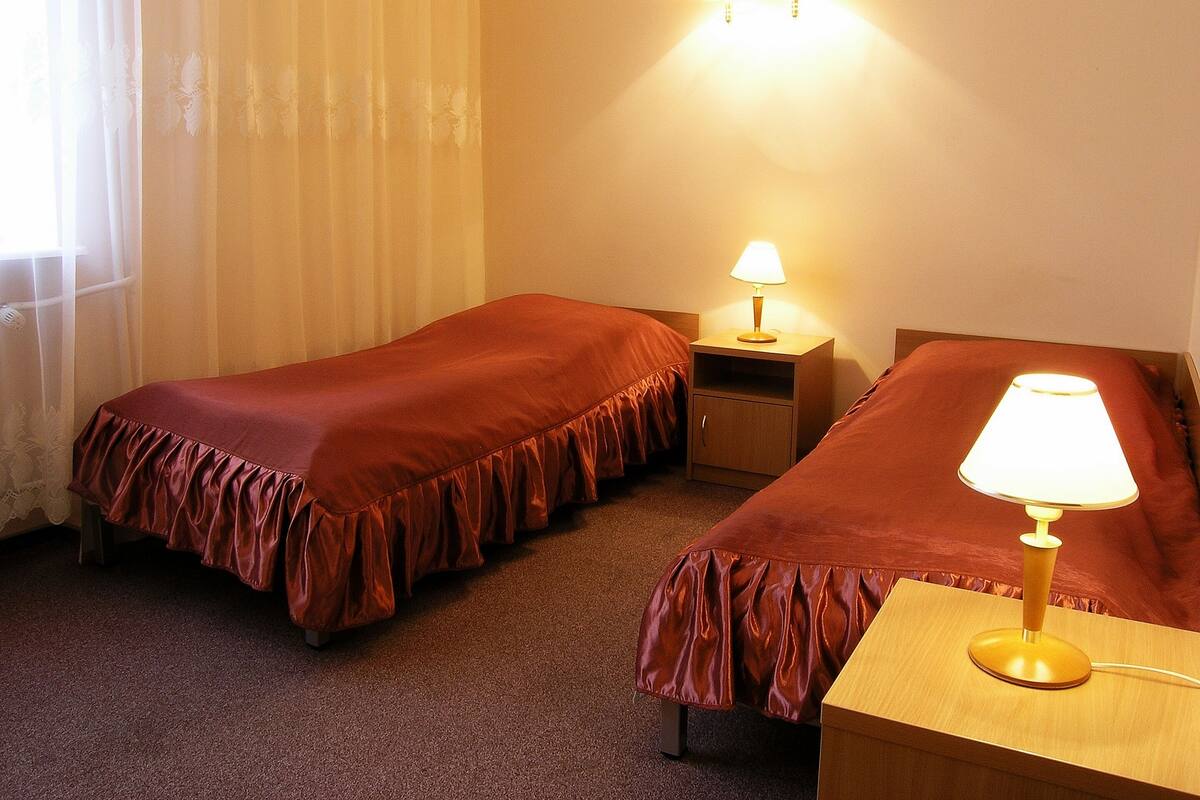 A hotel room with two beds, a nightstand with a lamp, and a window with sheer curtains.
