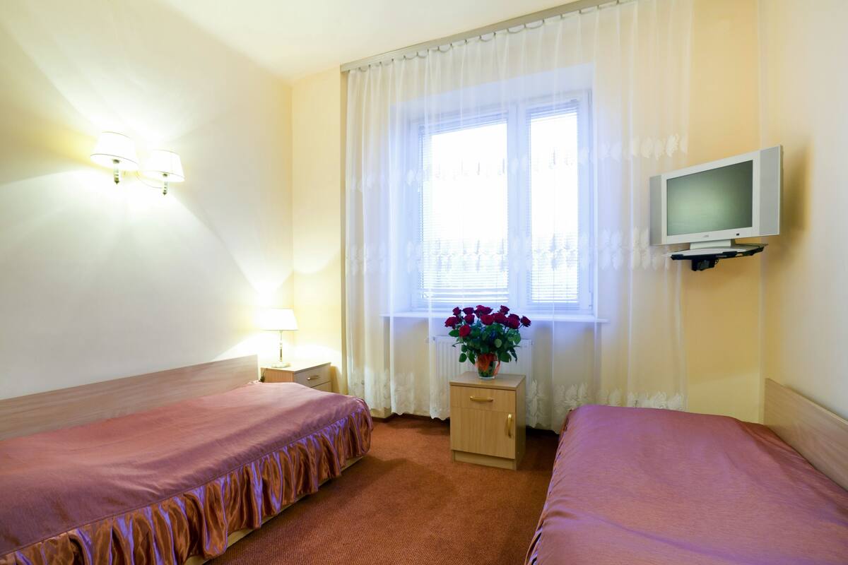 A hotel room with two beds, a TV, a nightstand, a lamp, and a window with curtains.
