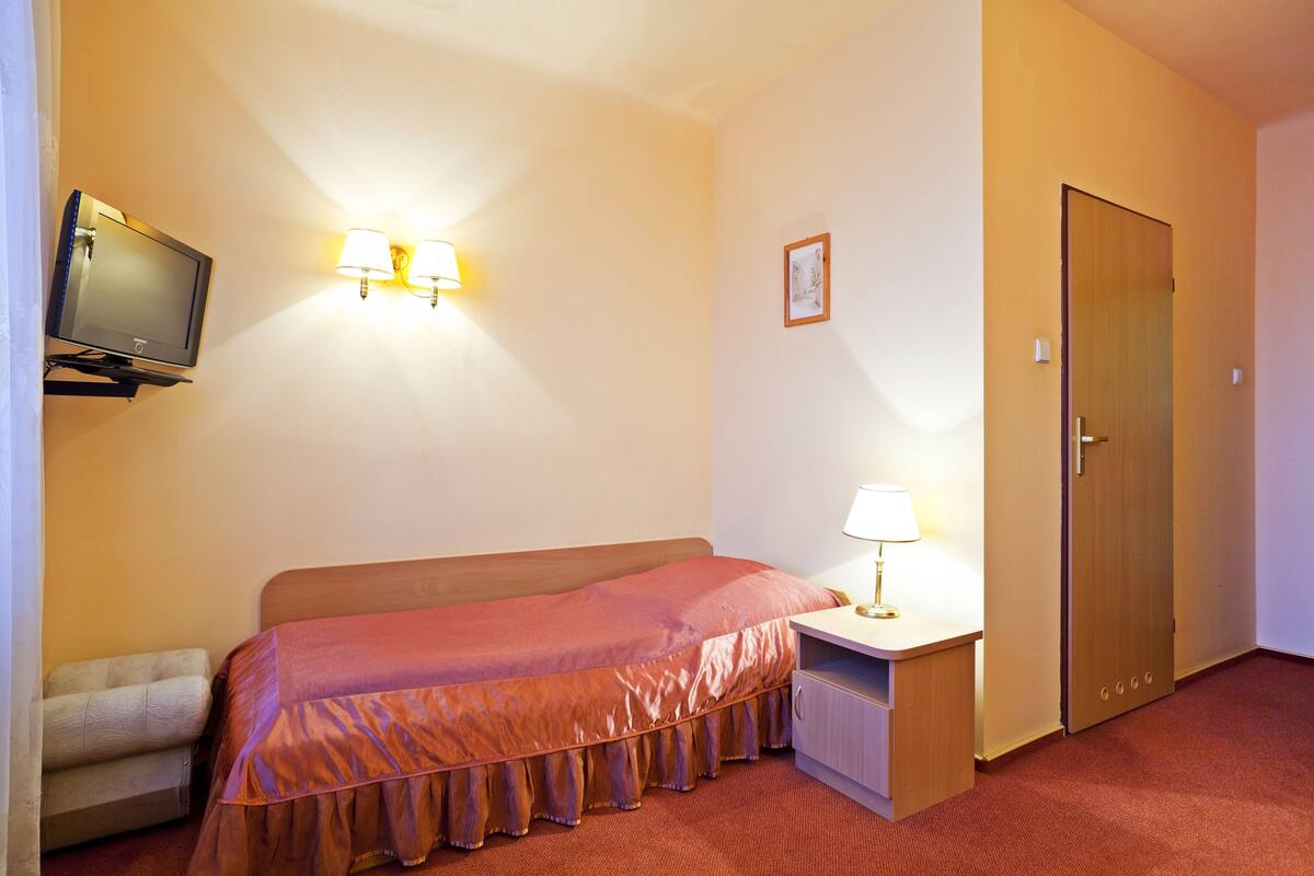 A hotel room with a bed, a TV, a desk with a lamp, and a door.