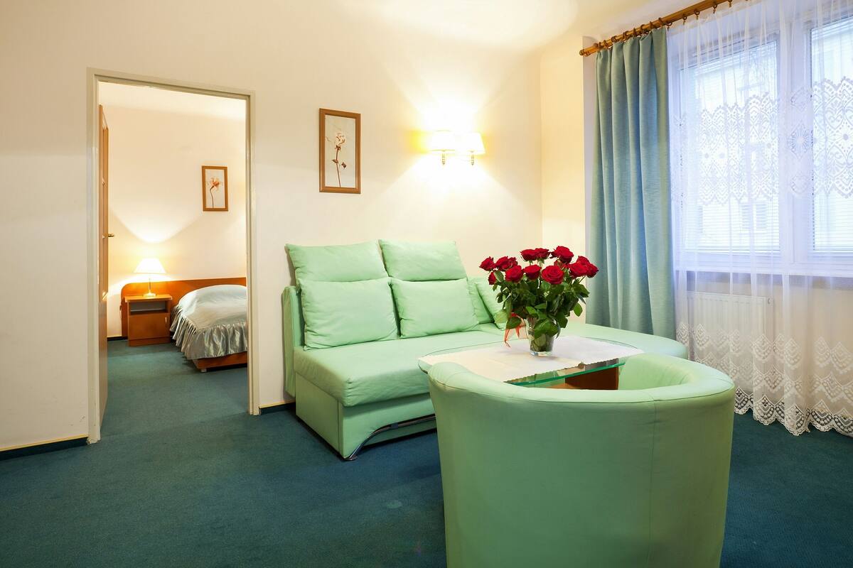 A room with a green sofa, a small table with a vase of red flowers, a bed with a wooden headboard, and a window with sheer curtains.