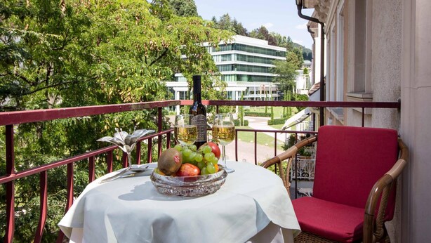 A balcony with a table set with fruit and wine glasses, a wicker chair, and a view of a building and trees. - Hotel Beek (Baden-Baden)