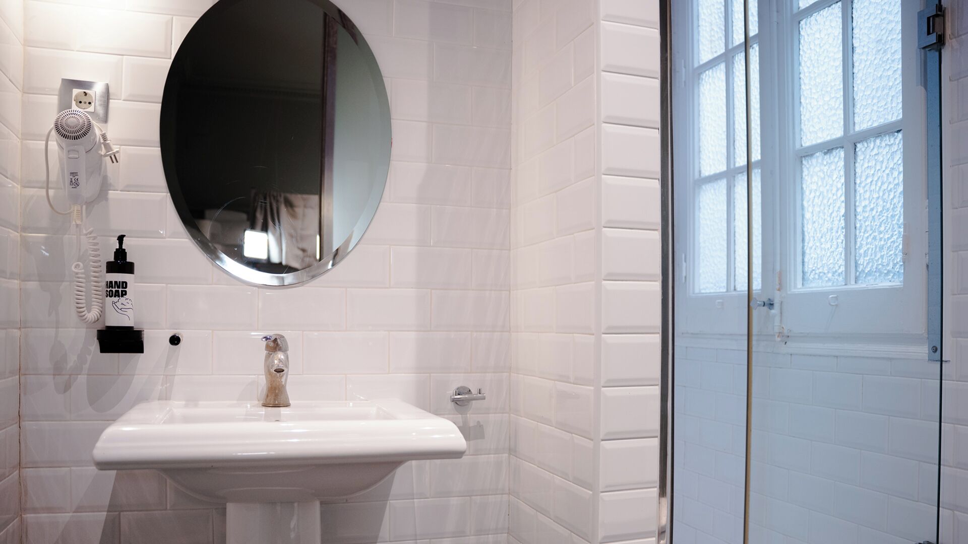 A bathroom with a white sink, a round mirror, a handheld soap dispenser, and a hairdryer on the wall.