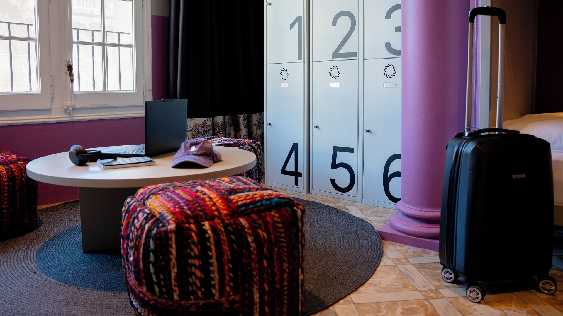 Une chambre d’hôtel avec une table ronde, un ordinateur portable, une valise et des poufs colorés.