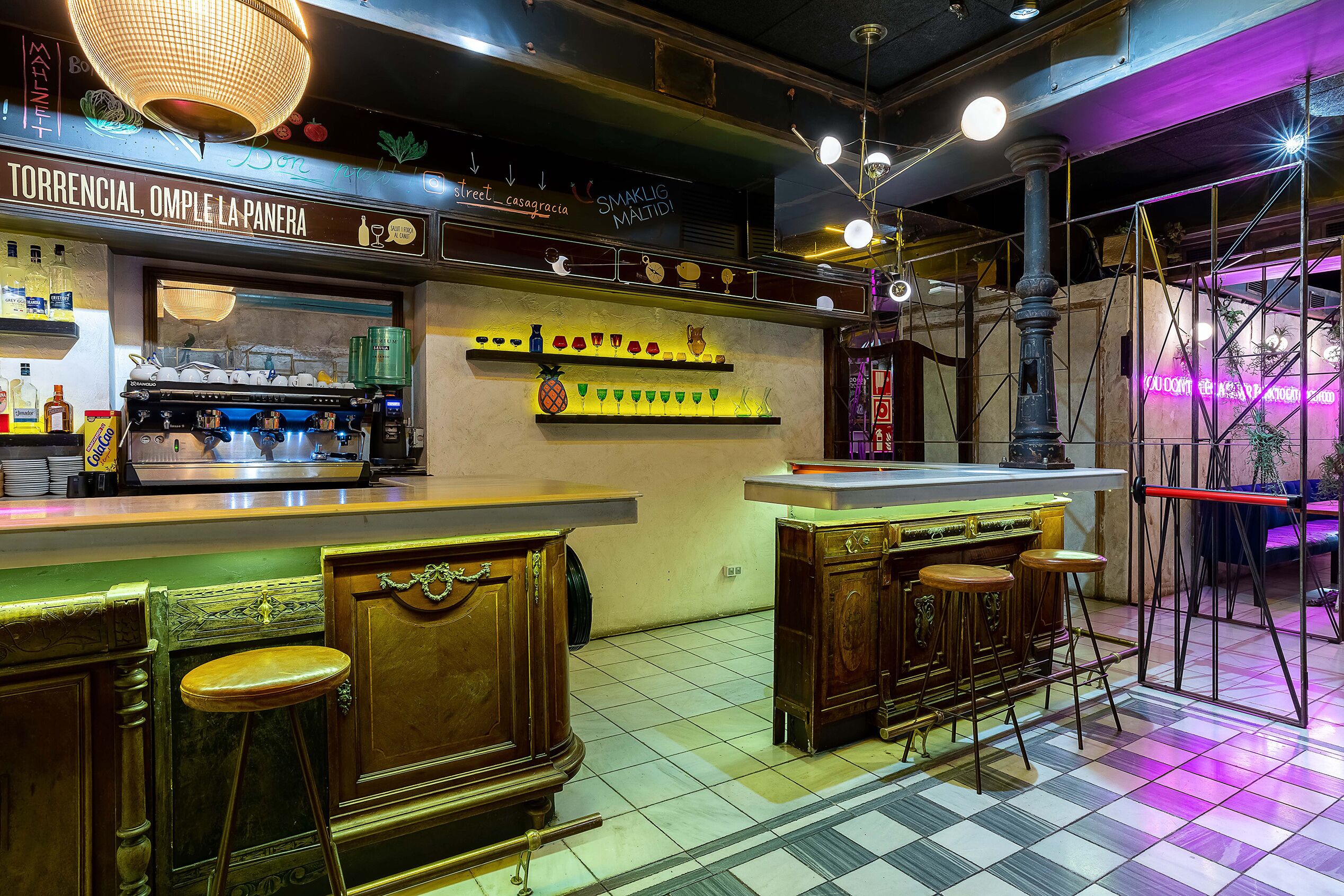 Una zona de bar con mostrador, taburetes y estantes con botellas y vasos.