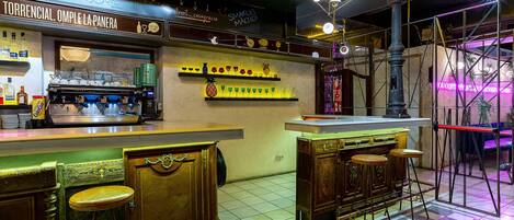 Una zona de bar con mostrador, taburetes y estantes con botellas y vasos.