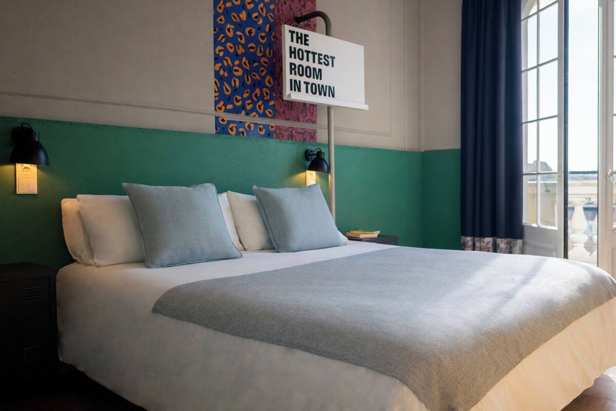 Un grand lit avec une couette grise et blanche, deux lampes de chevet et une pancarte sur le mur indiquant : « LA CHAMBRE LA PLUS BRANCHÉE DE LA VILLE ».