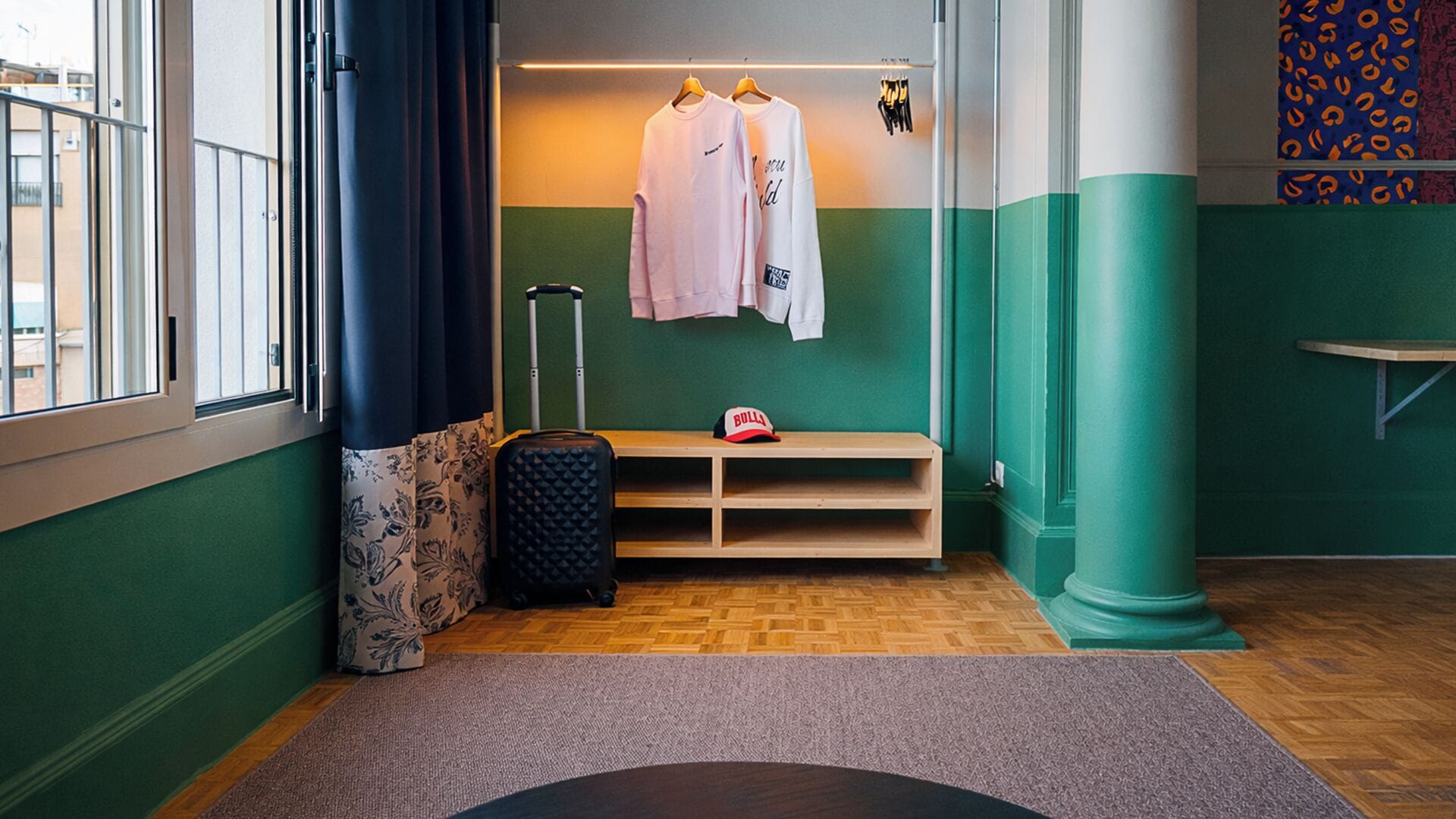 Una habitación con ventana, una maleta, un banco y una pared con dos camisas colgadas.