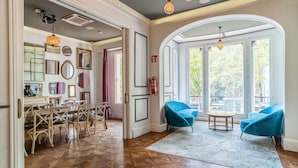 A spacious room with a dining area, two blue armchairs, a round table, and large windows with natural light.
