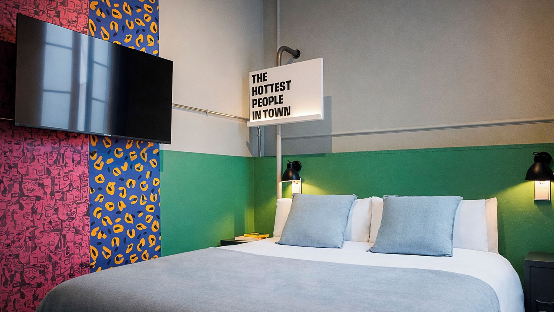 A hotel room with a bed, a TV mounted on the wall, and a sign that reads "THE HOTTEST PEOPLE IN TOWN".