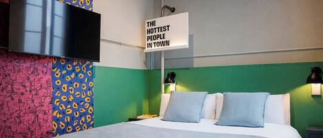 A hotel room with a bed, a TV mounted on the wall, and a sign that reads "THE HOTTEST PEOPLE IN TOWN".