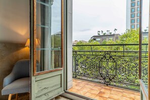 Un balcón con barandilla de hierro forjado, una silla y vistas a edificios y árboles.