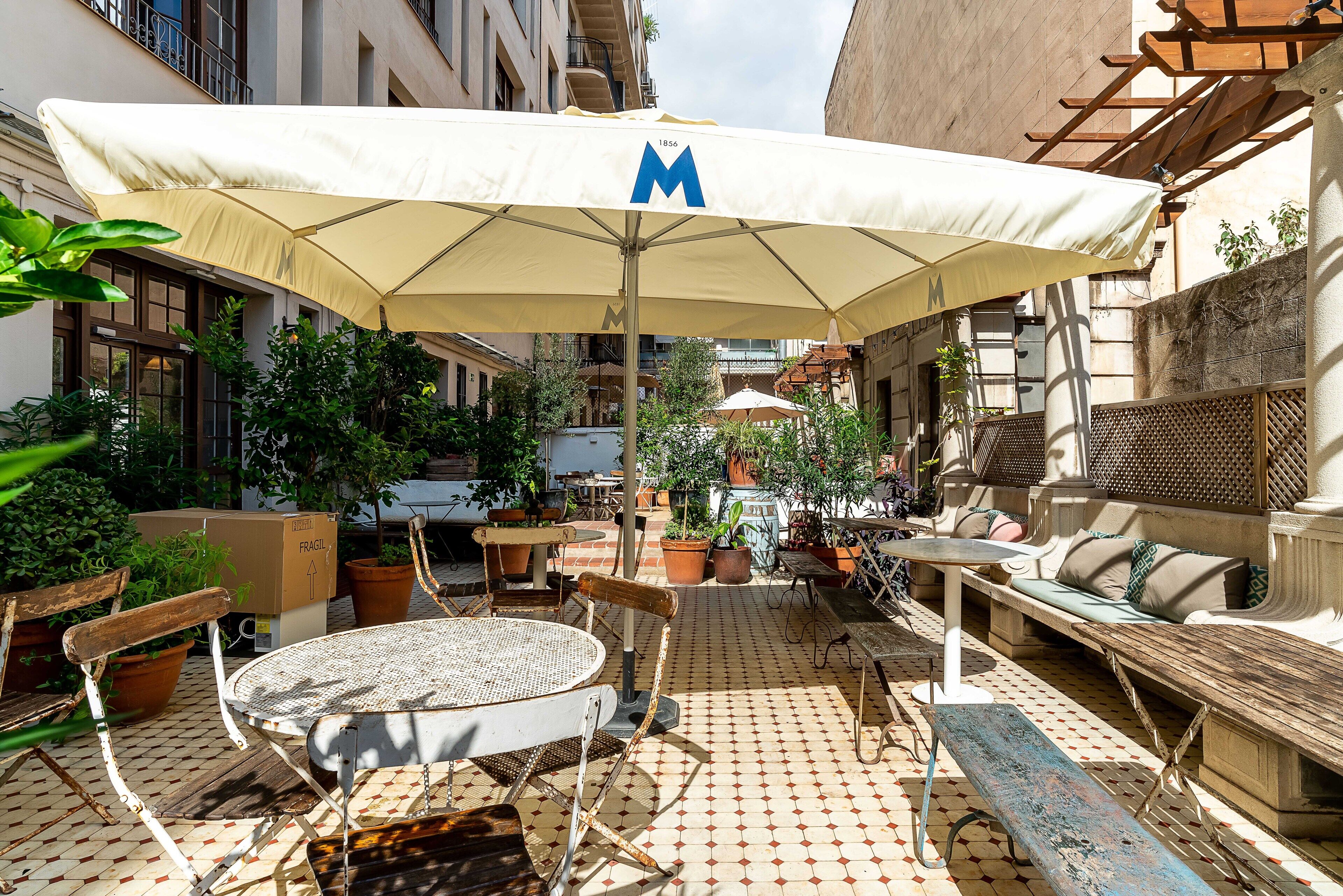 An outdoor patio with a round table, chairs, a bench, and an umbrella with a logo.
