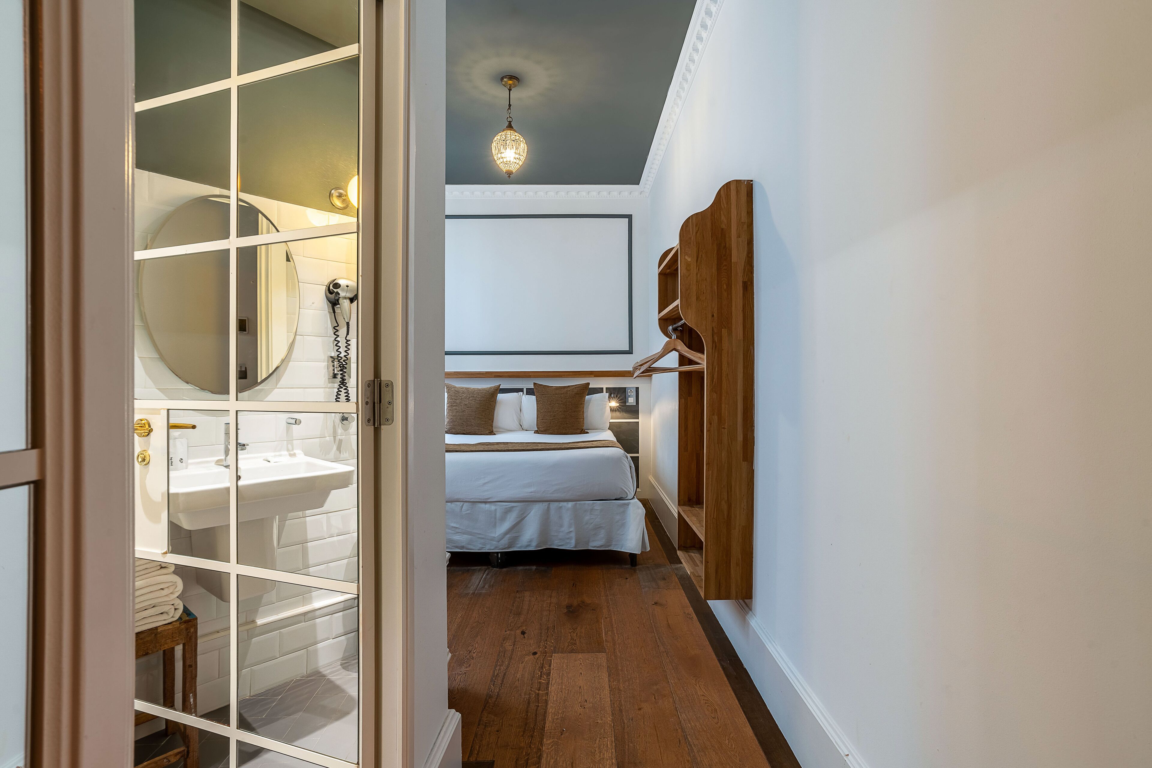 Quarto de hotel com piso de madeira, cama branca, guarda-roupa de madeira, espelho redondo e pia branca.