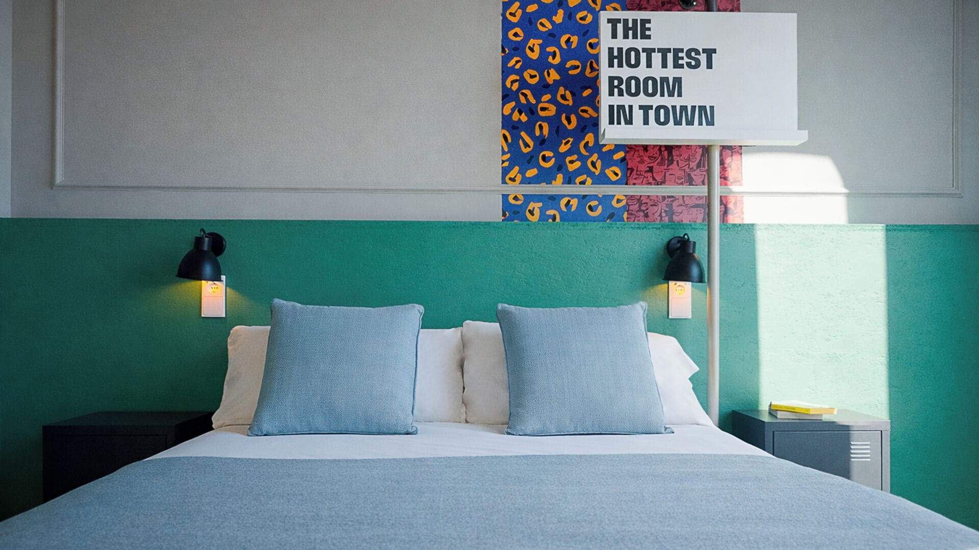 A hotel room with a bed, two pillows, two bedside lamps, a nightstand, and a wall sign stating "THE HOTTEST ROOM IN TOWN".