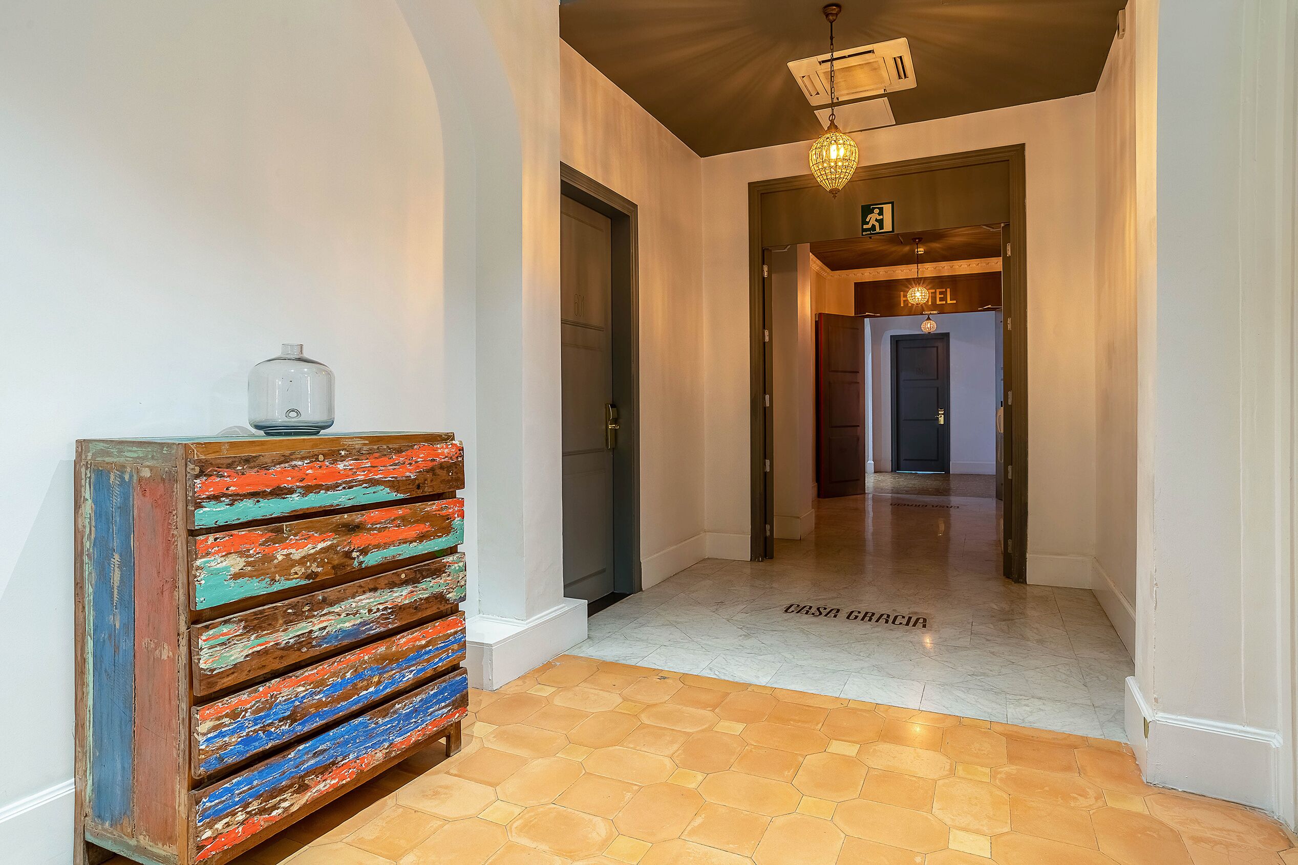 Un couloir d’hôtel avec une commode en bois, un sol carrelé et un panneau indiquant la direction des chambres.