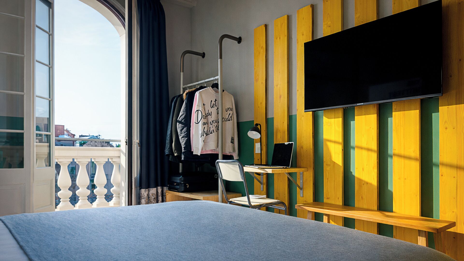 Quarto de hotel com cama, escrivaninha, cadeira, televisão e janela com vista para edifícios.