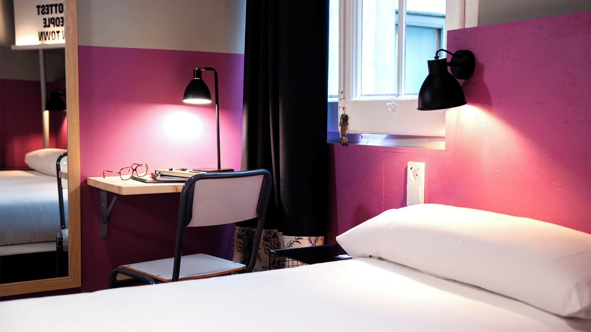 A hotel room with a bed, a desk with a chair, a mirror, and two wall-mounted lamps.