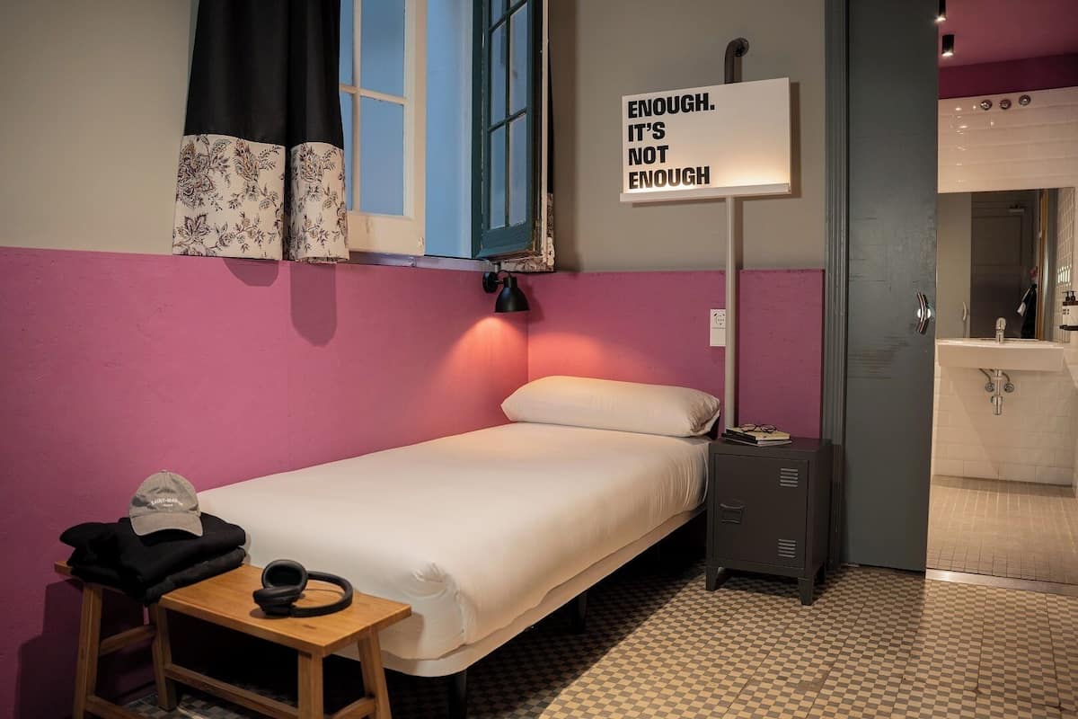 Une petite chambre à coucher, bien rangée, avec un lit, une table de chevet sur laquelle se trouvent une tasse et un chapeau, une commode, un panneau fixé au mur, une fenêtre avec des rideaux et une salle de bain visible à travers une porte ouverte.