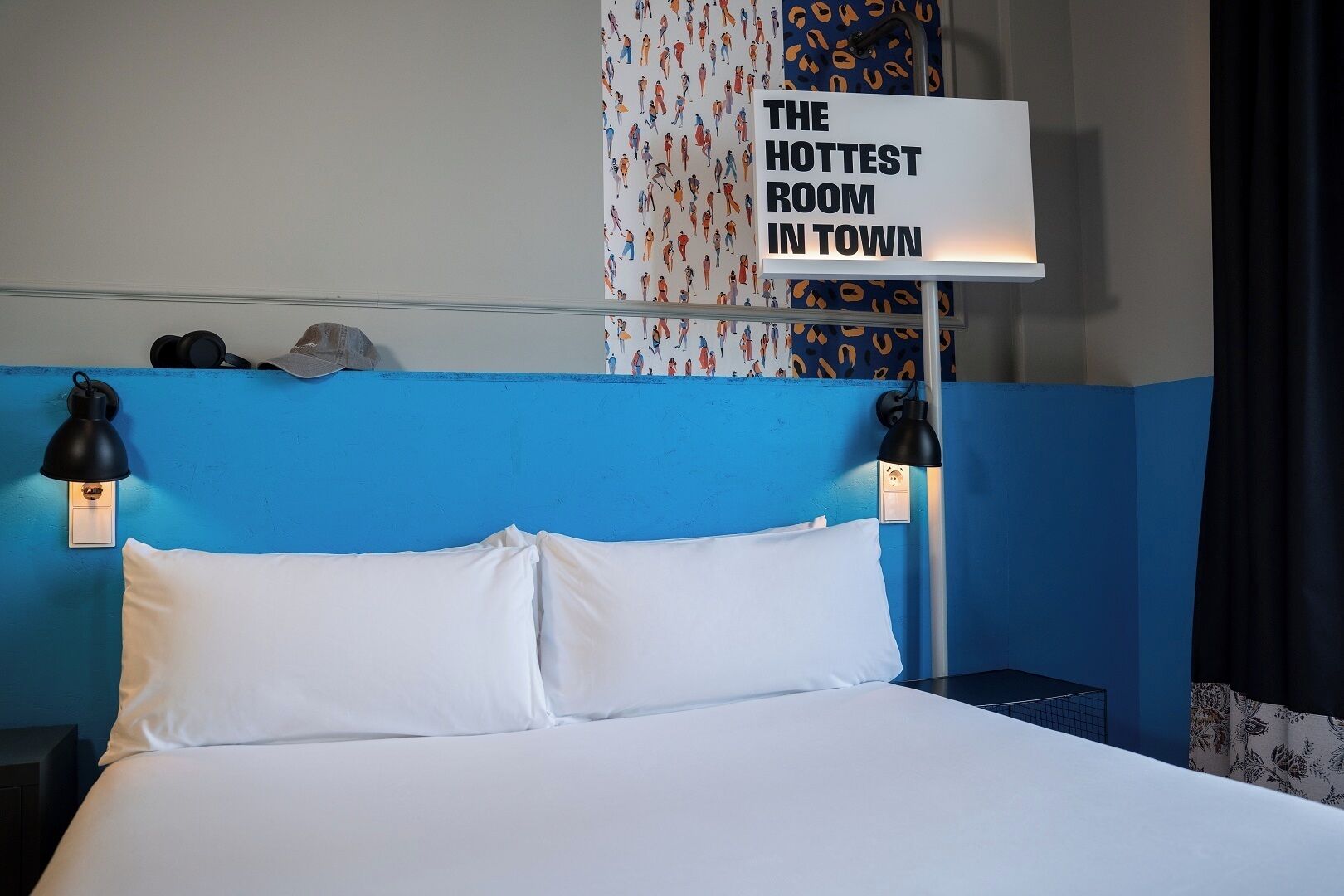 A hotel room with a blue accent wall, a bed with white linens, and two wall-mounted lamps. A sign above the bed reads "THE HOTTEST ROOM IN TOWN".