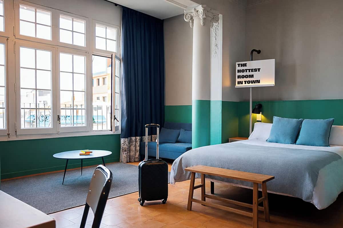 Une chambre d’hôtel avec un lit, un bureau, une chaise, une valise et une fenêtre avec des rideaux.