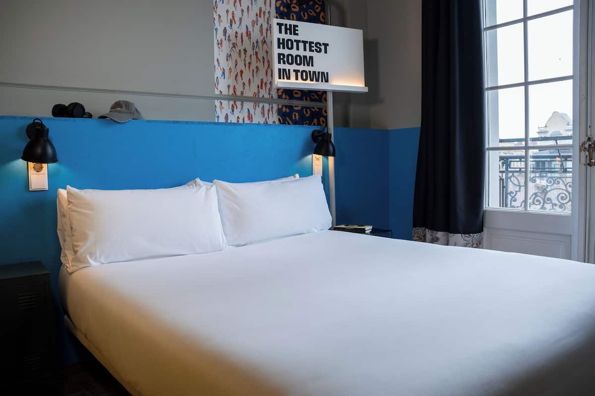 Une chambre d’hôtel avec un grand lit, deux lampes de chevet, une pancarte indiquant « LA CHAMBRE LA PLUS BRANCHÉE DE LA VILLE » et une fenêtre donnant sur un balcon.